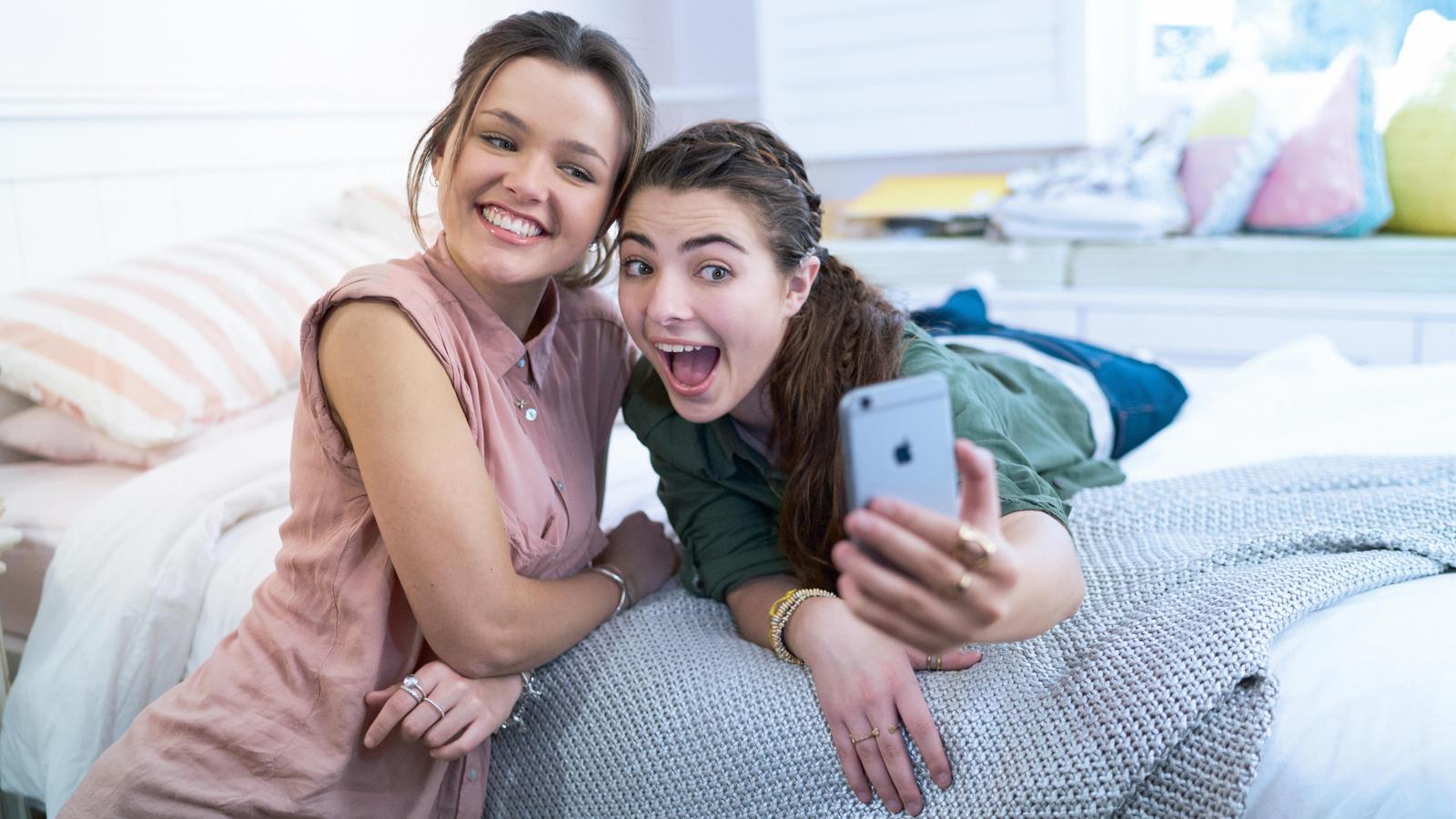 Two friends pose for a photo with an iPhone in a bedroom