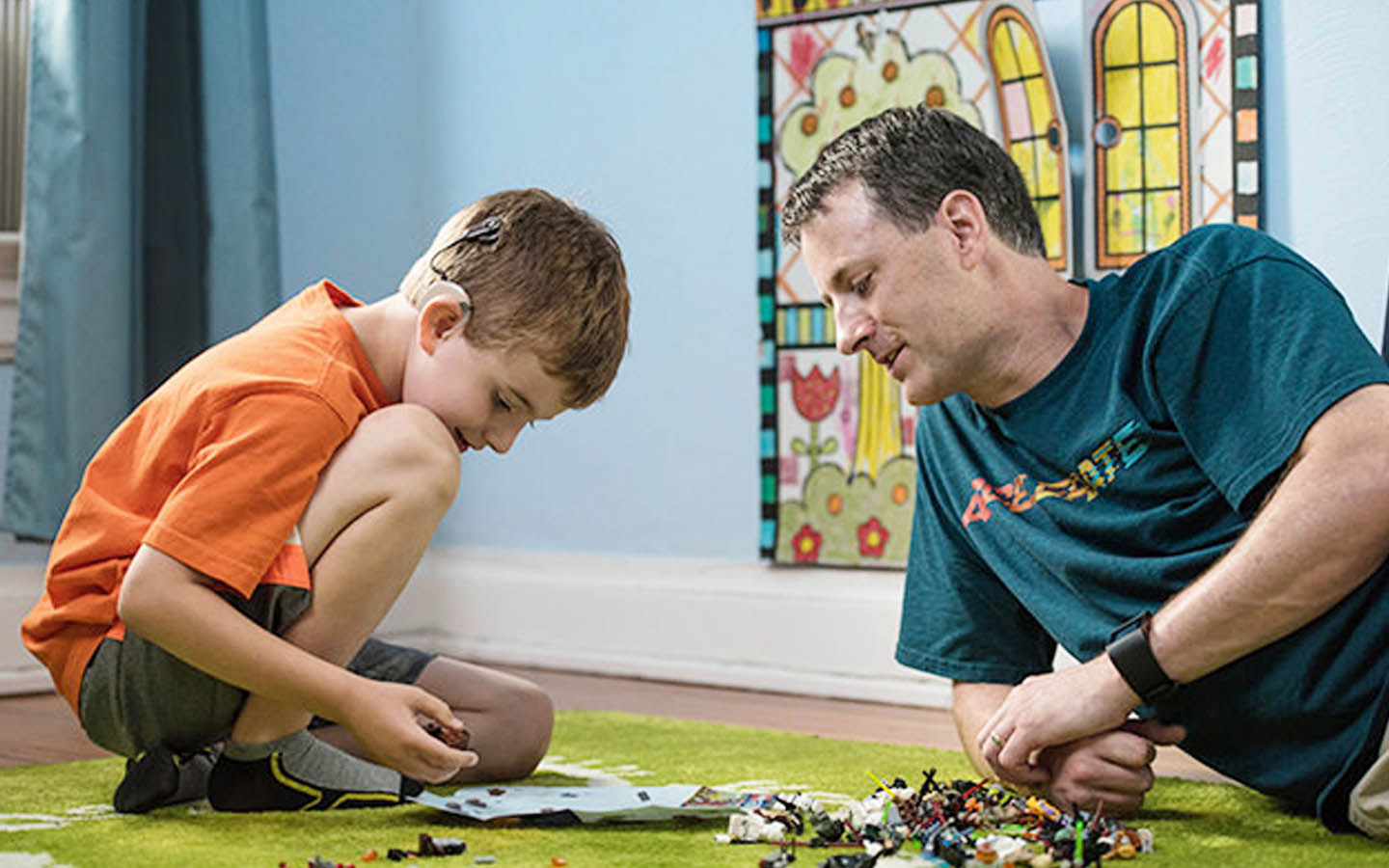A boy with a Cochlear implant plays on the floor as his dad watches on