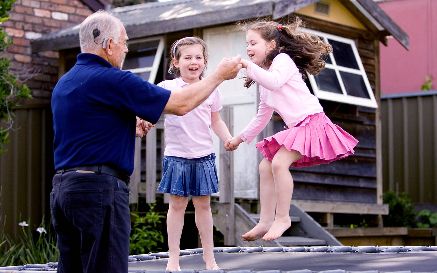 A Cochlear recipient holds his two grandaughters' hands as they jump on a trampoline