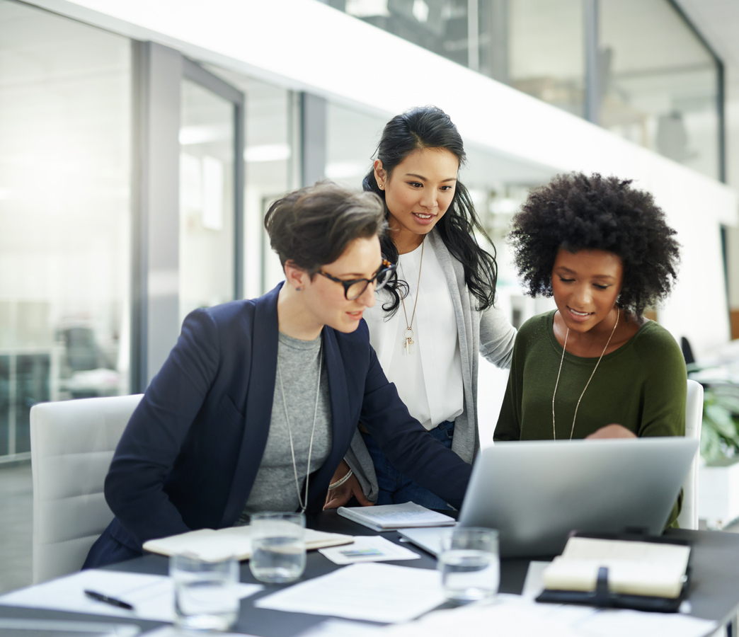 Three professional women, privacy experts, in business attire gathered around a laptop, engaged in a focused discussion about how privacy is considered in their project.